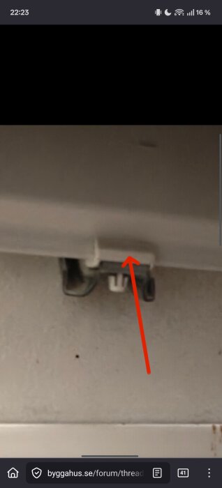 En vit plastkrok med en röd pil som pekar på den, monterad mot en grå yta.