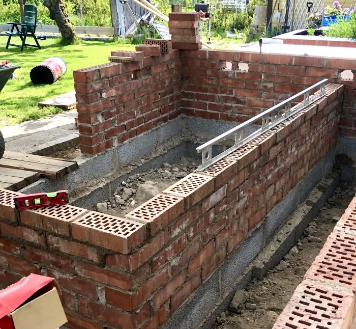 Mur av röda tegelstenar byggs på leca-block och betongsula, med vattenpass ovanpå, i en utgrävd trädgårdsmiljö.