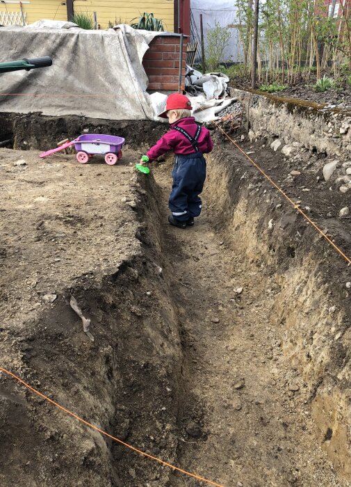 Barn i byggkläder leker nära en utgrävd grop i trädgården, med en liten rosa vagn och leksaksspade.