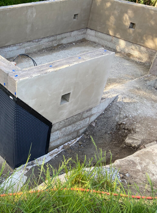 Grundläggning för växthus med murade väggar och fyrkantiga utskärningar, omgiven av jord och gräspartier.