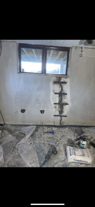 Vägg med lagade hål och sprickor under fönster, armering synlig, slipverktyg och cementmaterial på golvet.
