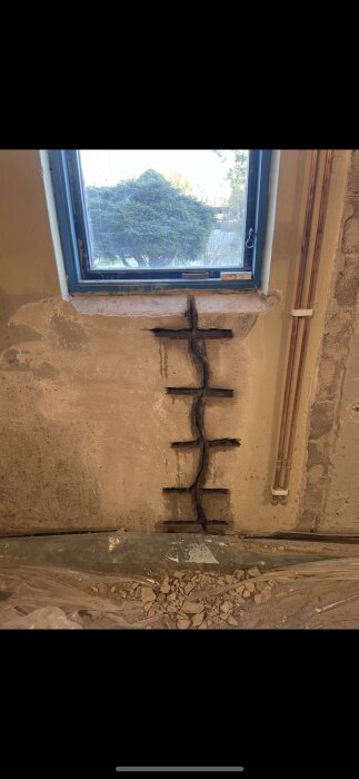 Sprucken betongvägg under ett fönster, med sågade spår och plast över golvet. Rör syns på höger sida av väggen.