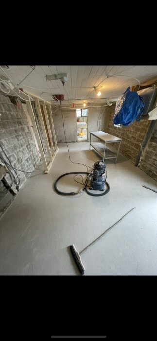 Renoveringsrum med slipdammssugare, nyputsade väggar och elledningar. En arbetsbänk och en sopborste ses på betonggolvet.