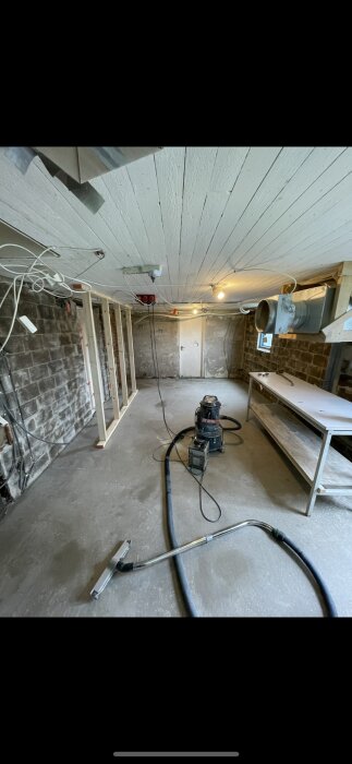 Renoveringsprojekt med verktyg och material i ett rum med obehandlade väggar. En dammsugare med lång slang syns på golvet.