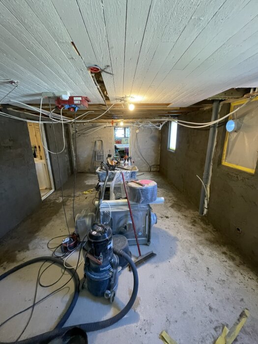 Renovering av rum med dammsugare, byggmaterial och verktyg på golvet, oskyddade väggar och vitmålat tak, elektriska ledningar synliga i taket.