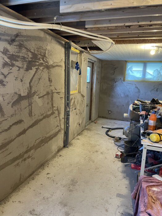 En källare under renovering med putsade väggar och verktyg på arbetsbänkar. Två fönster är täckta med plast.