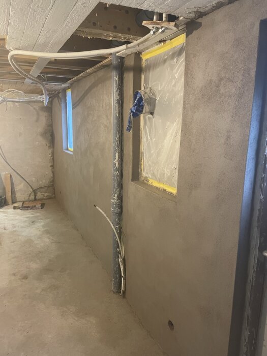 En putsad vägg med rör och ett täckt fönster under renovering i en källare.