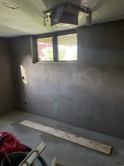Vägg i ett rum under renovering, nyslammat med putsbruk, fönster med utsikt och material på golvet.
