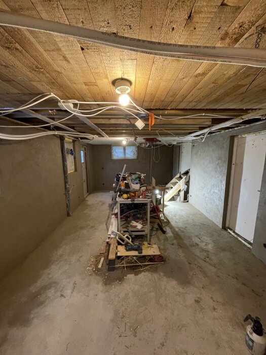 Renoveringsprojekt med oputsade väggar, trätak, arbetsbänk full av verktyg och material, taklampor och kablar i ett halvfärdigt rum.