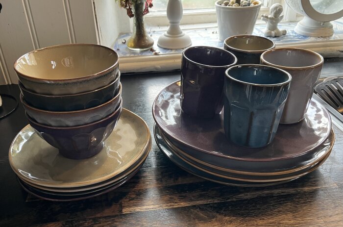 Staplade keramikskålar och muggar i olika färger på tallrikar vid ett fönster.