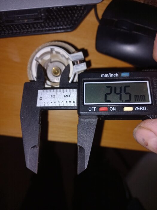 Radiatortermostat av stor diameter mäts med digital skjutmått som visar 24.5 mm, placerad på ett skrivbord intill en datormus.