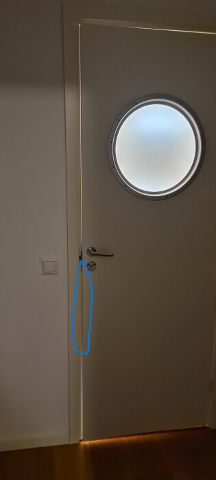 Innerdörr med glipande springa vid dörrkarmen, markerad med blå linje, nära ett runt fönster och dörrhandtag.