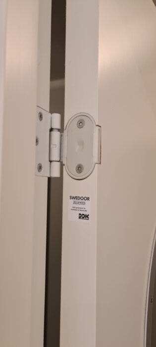 Gångjärn på vit innerdörr med märkningen Swedoor. Dörren har problem med att stänga, då den slår i dörrkarmen vid låset.