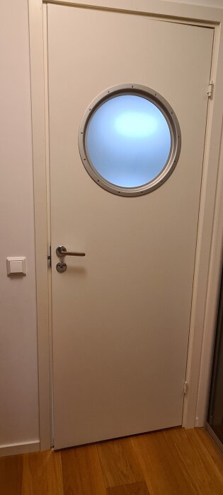 Vit innerdörr med runt fönster och handtag, öppnas mot en tröskel och visar dörrkarmen där problem kan uppstå vid låset.