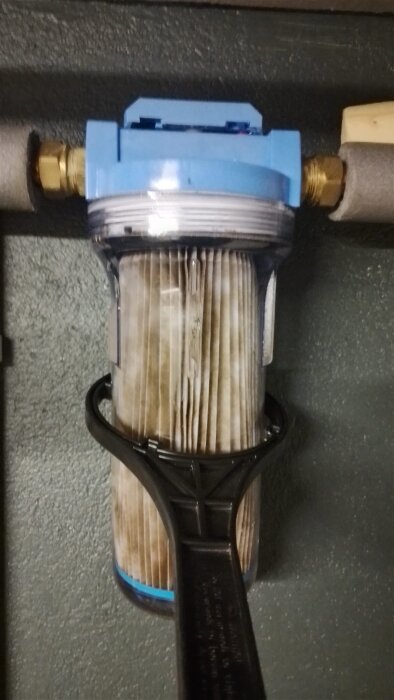 Vattenfilter inskruvat i ett rörsystem, med ett plastverktyg som försöker lossa det. Filtret verkar fast och är delvis skadat.