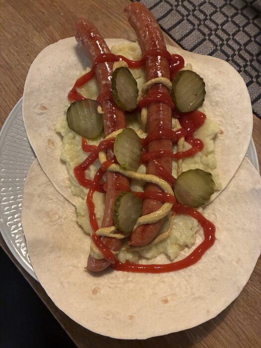 Två varmkorvar med ketchup, senap, skivad gurka och potatismos på tortilla.