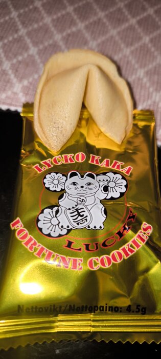 Lyckokaka förpackad i en guldfärgad påse med en tecknad katt, märkt "Lycko Kaka Lucky Fortune Cookies". En lyckokaka ligger ovanpå.