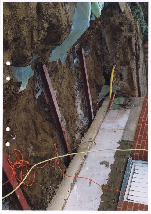 Metallbalkar stöder ett schakt vid husgrund med synliga kablar, efter förstärkning mot markrörelser.