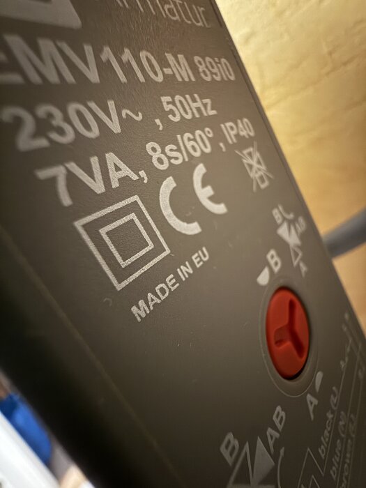 Närbild på en elkomponent märkt med specifikationer som 230V, 50Hz, och CE-märkning, med en röd ratt i mitten.