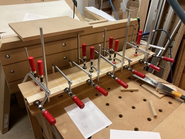 Arbetsbänk i verkstad med flera limtvingar fästa på brädor och en sågad mdf-bit, verktyg och projektplaner på bordet.