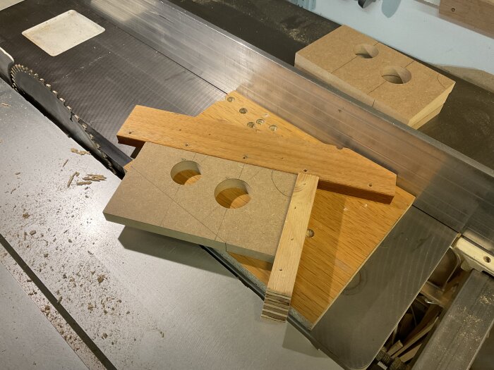 Sågverkstad med MDF-bitar och träjiggar, två cirkulära hål i MDF-skivor, placerade på en arbetsbänk nära en sågklinga.