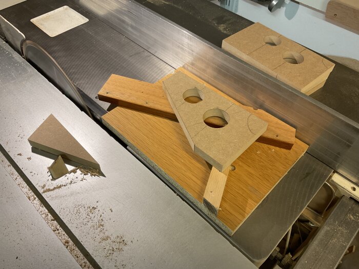 MDF-bitar med runda hål och triangulära delar på en arbetsbänk, omgivna av sågspån. En del av ett snickeriprojekt i verkstaden.