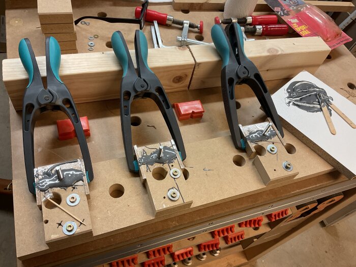 Fyra klämmor håller samman bitar av MDF med epoxi och skruvar på en arbetsbänk i verkstad. Verktyg och material syns i bakgrunden.