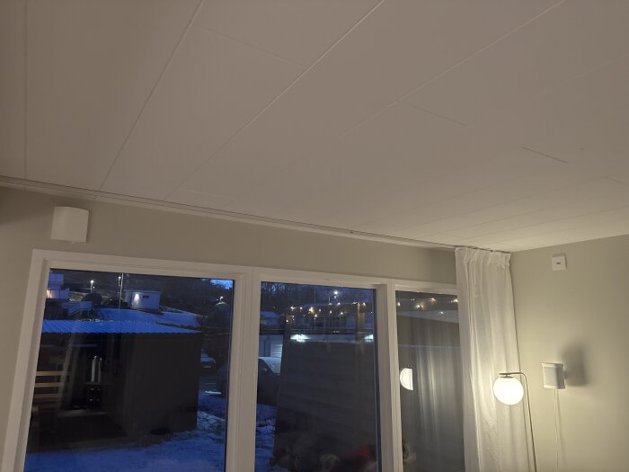 Vardagsrum med ljusa väggar, gardinskenor, vit gardin, en golvlampa och fönster mot en utomhusmiljö med snö och byggnader.