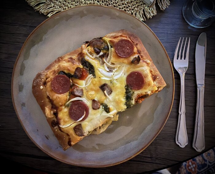 Närbild på en bit pizza toppad med salami, fläskfilé, lök, pesto, tomat och ost, serverad på en tallrik med bestick bredvid.