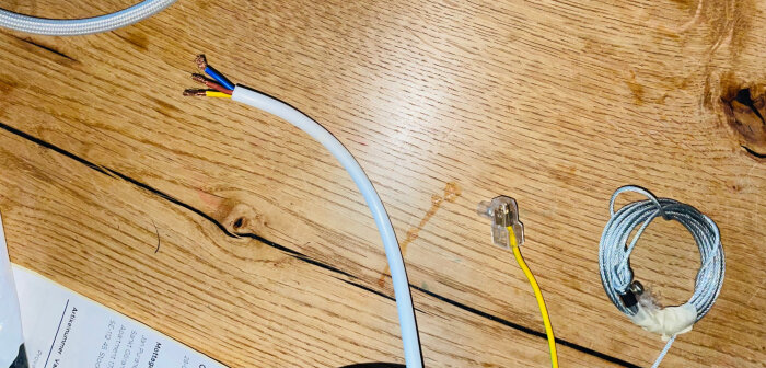 Elkabel med exponerade ledare i brun, blå och gulgrön, en gul kabel i klämma och en rulle ståltråd på träbord.