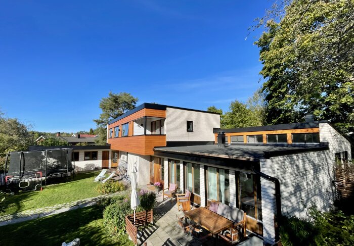 Arkitektritad villa med ny fasad i liggande furupanel och teakdetaljer, omgiven av grönska, utemöbler och en studsmatta i trädgården.