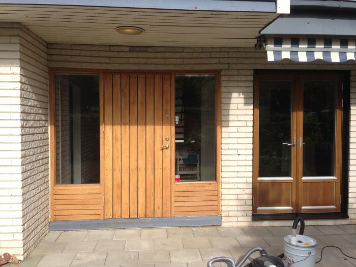 Entré med ljusbrun träpanel, mexitegelväggar och mörkblå/vit randig markis.