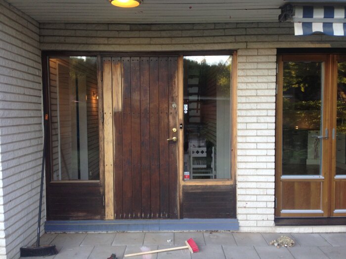 Entrédörr med mörk träfinish mellan vita tegelväggar, omgiven av stora fönster och en blåvitrandig markis ovanför.