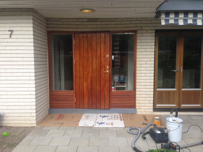 Entrédörr av trä med glaspartier omgiven av ljus mexitegelvägg. Verktyg och material på marken samt randig markis ovanför.