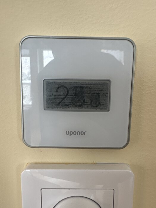 Termostat av märket Uponor monterad på en vägg, omgiven av en ljusgul bakgrund, visar 23,5 grader på displayen.