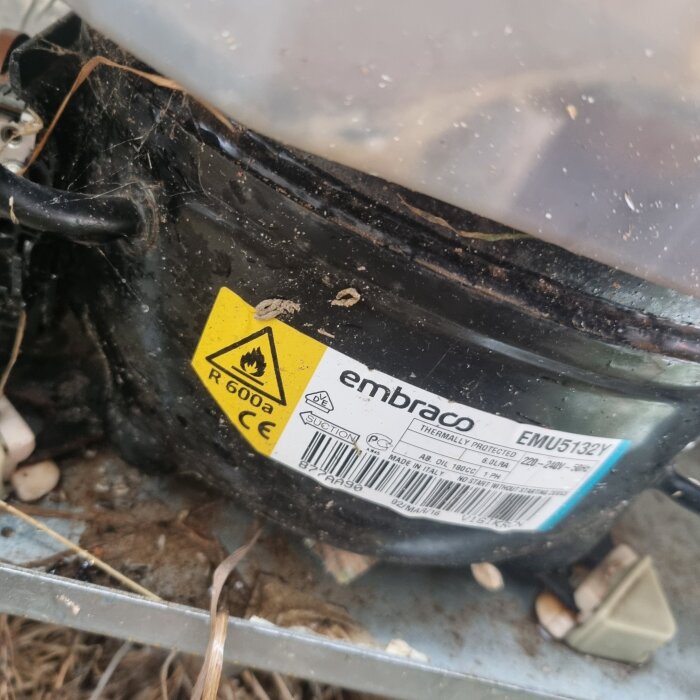 Etikett på en kompressor för värmepump, märkt med R600a och Embraco, omgiven av smuts och naturmaterial.