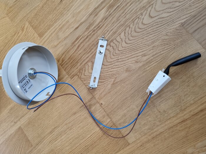 Ojordad lamputtagskomponent med blå och brun kabel, vit kopplingskåpa och metallbeslag ligger på trägolv.