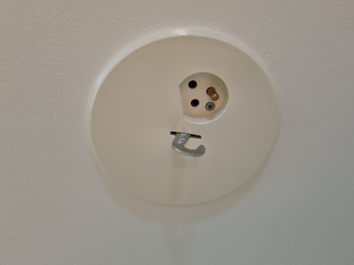 Gammalt jordat lamputtag i tak med krok för lampa och synlig skruv, monterat på vit vägg.