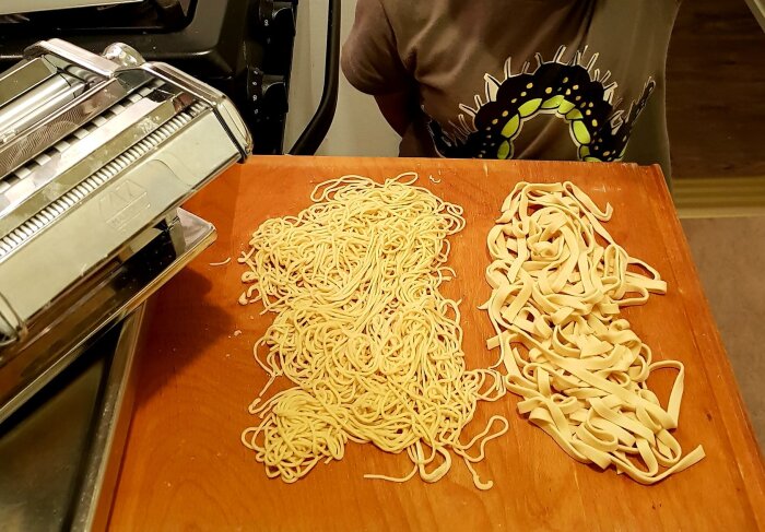 Hemgjord pasta bredvid en pastamaskin, med både smala och breda sorters pasta på ett träbord.