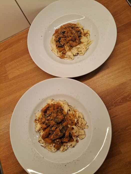 Två vita tallrikar med hemgjord pasta toppad med kött- och svampragu, dekorerad med spenatblad, på en träbänk.