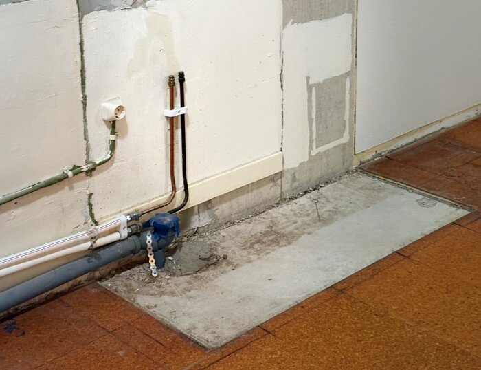 Vägganslutning med exponerat golv och rör, delvis borttagen golvbeläggning vid hörnet av ett rum, visar lager över betong.