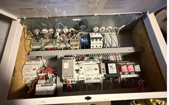 Öppen kontrollpanel för värmepump med kablar och komponenter, inkl. överklistrad etikett.