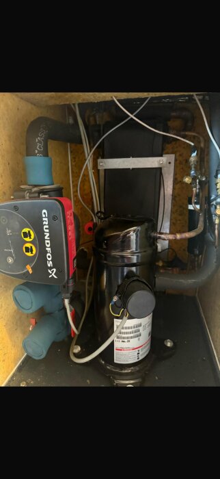 Inuti en värmepump från 2013, uppdaterad 2022 med ny kompressor och cirkpump, visas en Grundfos-pump och överklistrad etikett.