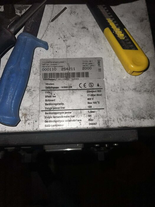 Bilden visar en pannetikett med specifikationer samt verktyg som en skruvmejsel och en stanskniv på en smutsig yta.