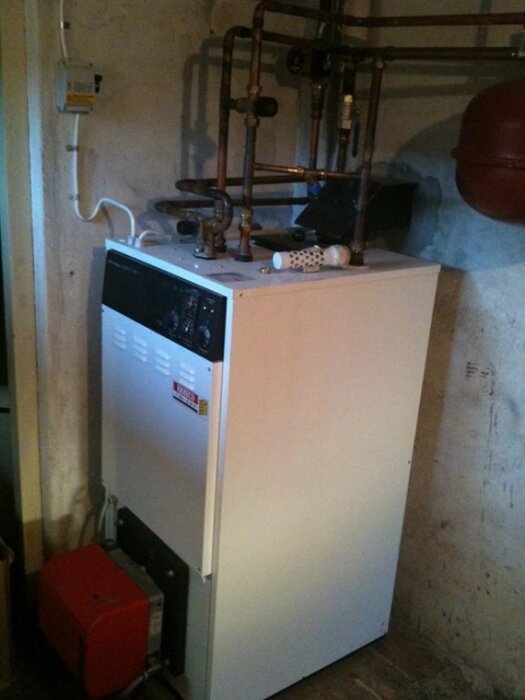 Pelletspanna med rördragning ovanpå, med kontrollpanel och vit plåt framför. Elpatronen finns bakom den vita plåten.