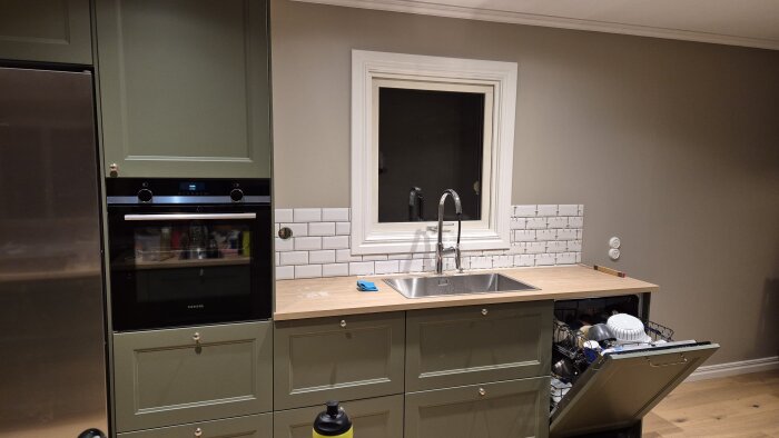 Grön köksinredning med målad skafferidörr, nykaklad vägg ovan diskbänk och öppen diskmaskin fylld med disk.