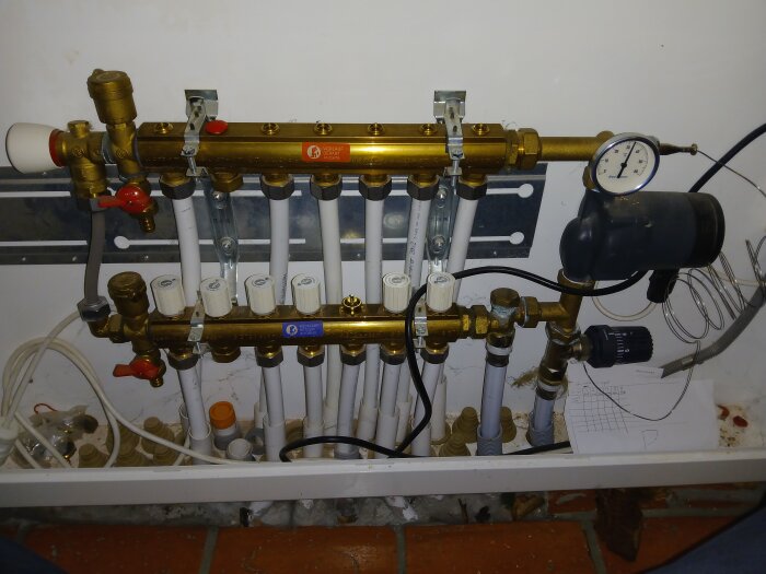 Rörkopplingar för golvvärme med manometer och ventilanslutningar, synlig T-skarv mellan tillopp och retur i ett hus med vattenburen golvvärme.