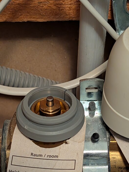 En ventilhylsa med en metallaxel och ett märklappar med texten "Raum/room" är monterad på en vägg med synliga kablar.