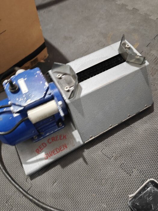 En maskin med en blå motor och texten "Red Creek Sweden" på sidan, placerad på ett golv av grått gummi.