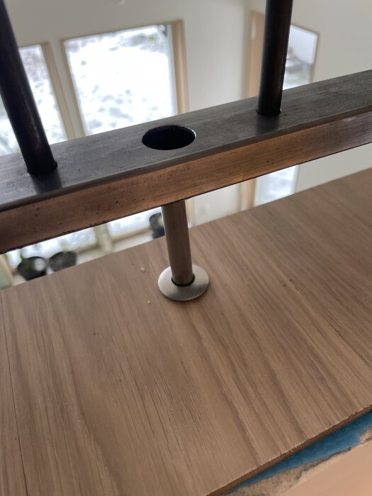 Närbild av räcke med metallcylinder monterad på träyta, del av byggprojekt, snöigt landskap syns genom fönstret i bakgrunden.